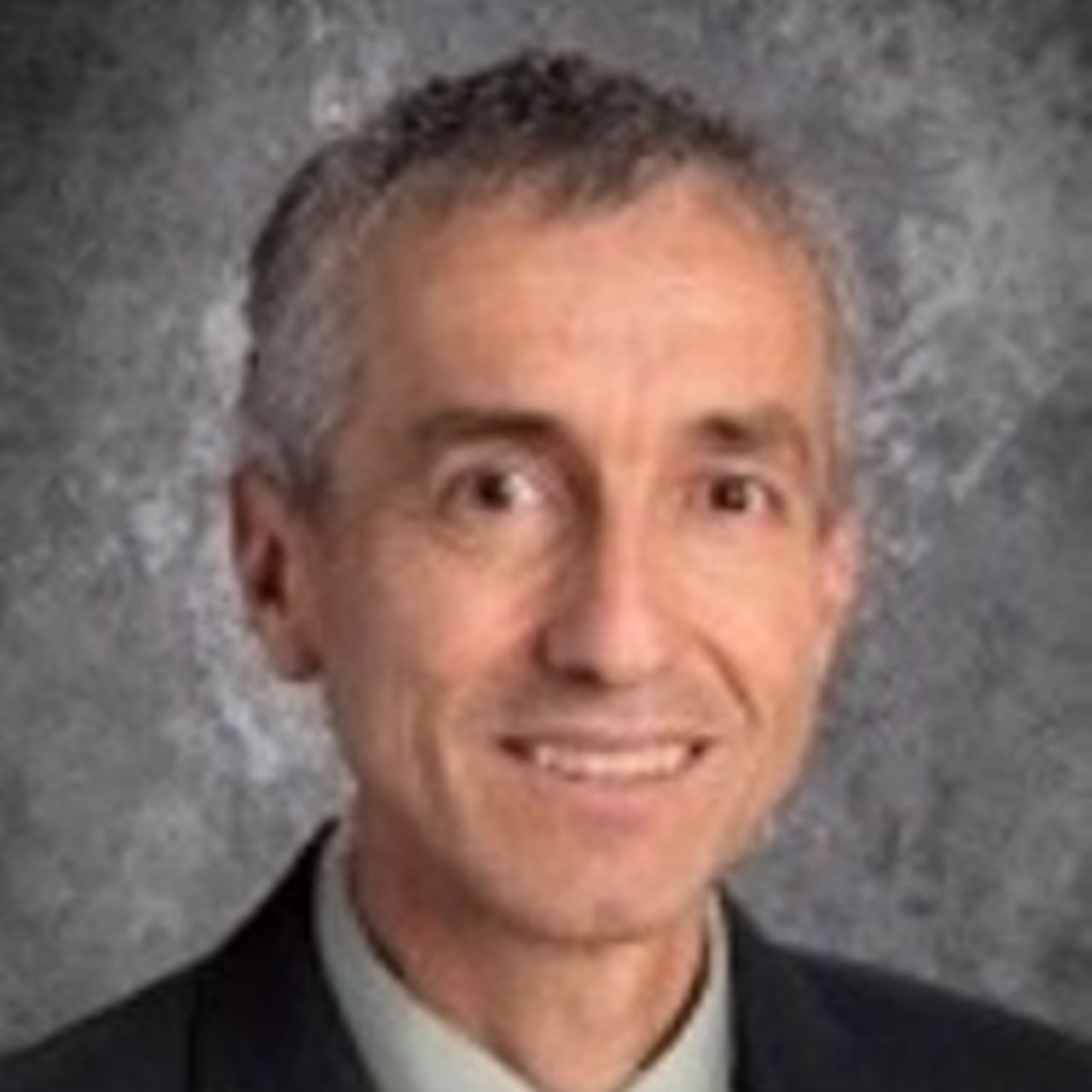 A person with short gray hair, wearing a suit and tie, smiling against a gray textured background