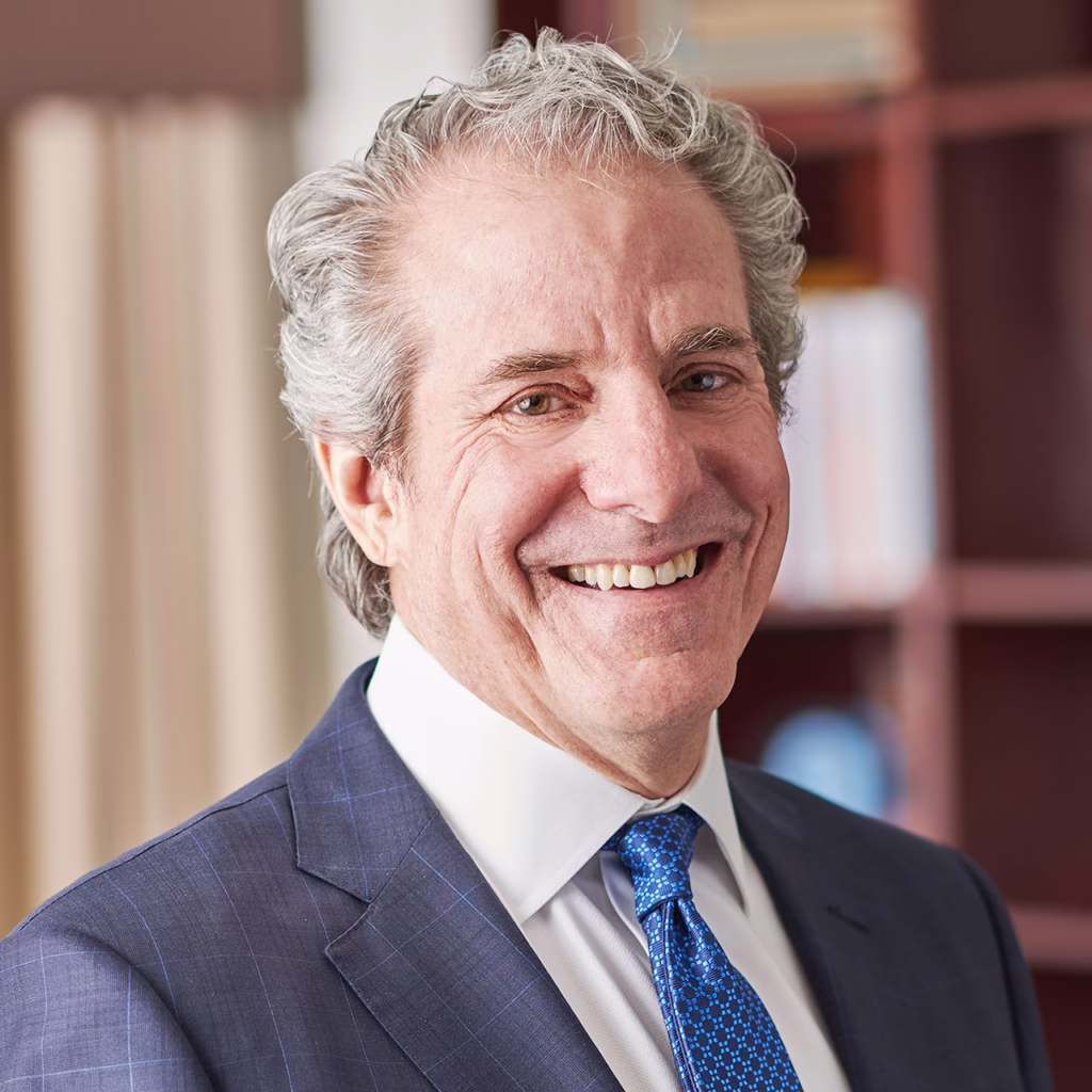 Smiling man in a suit with a blue tie, standing in an office setting with bookshelves in the background