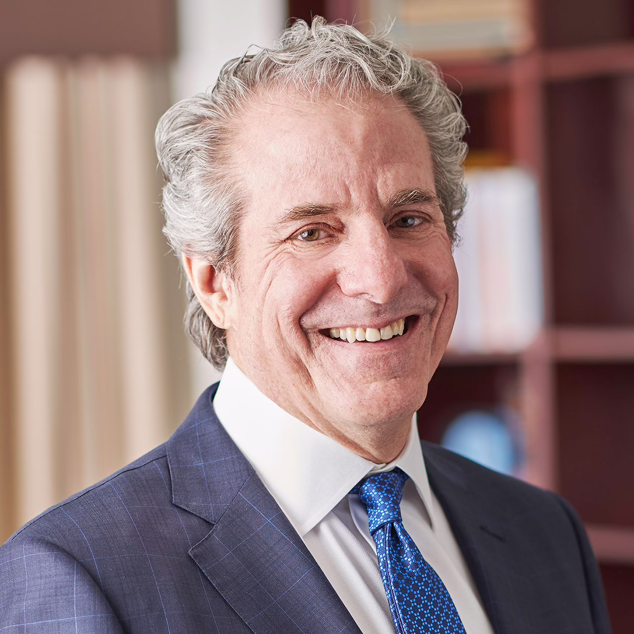 Smiling man in a suit with a blue tie, standing in an office setting with bookshelves in the background