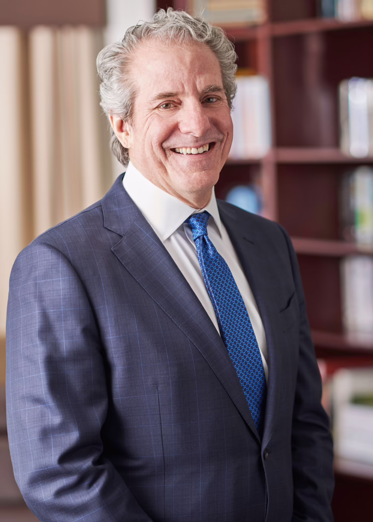 Smiling man in a suit and blue tie stands in an office with bookshelves in the background