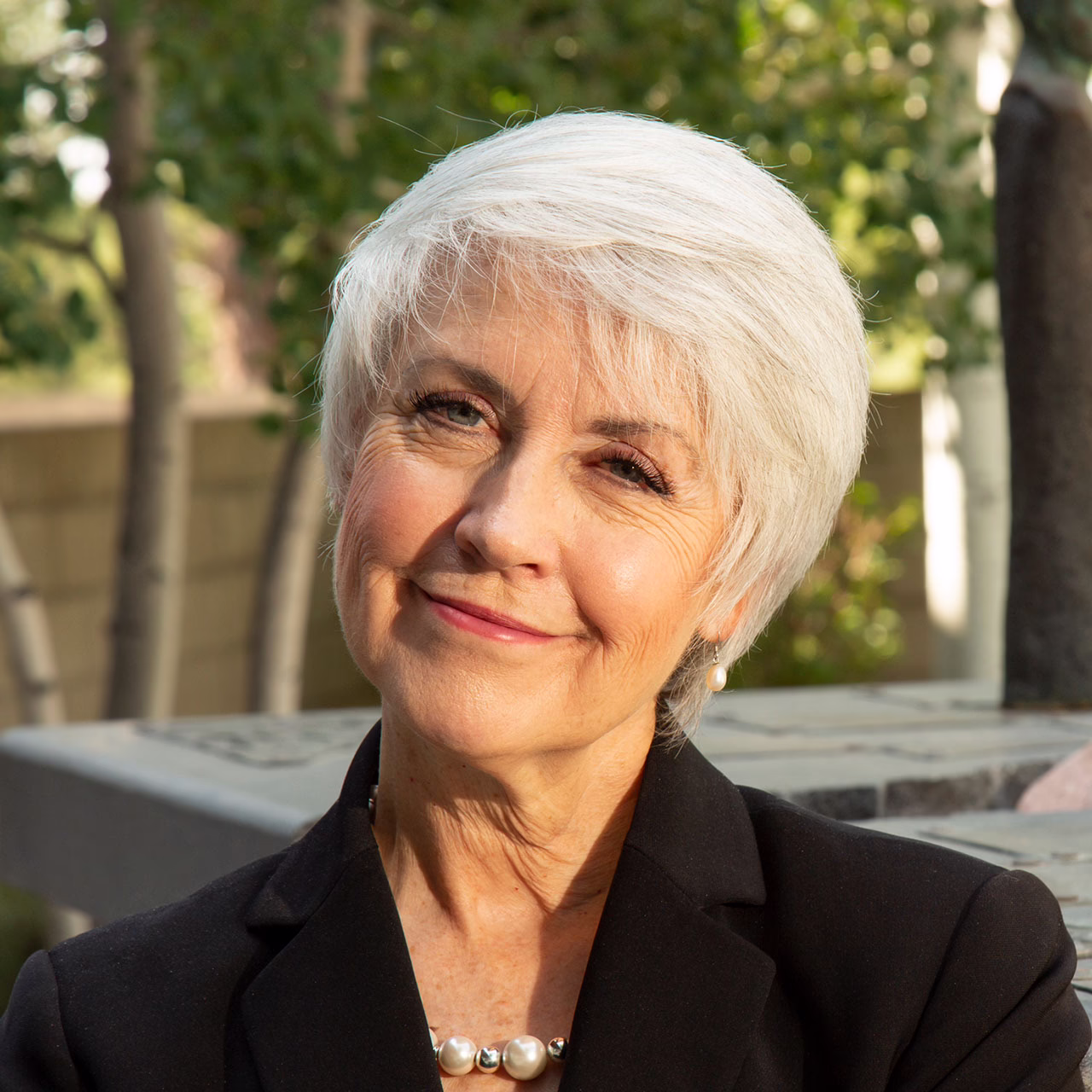 A woman with short white hair smiles, wearing a black blazer and pearl necklace, outdoors with trees in the background