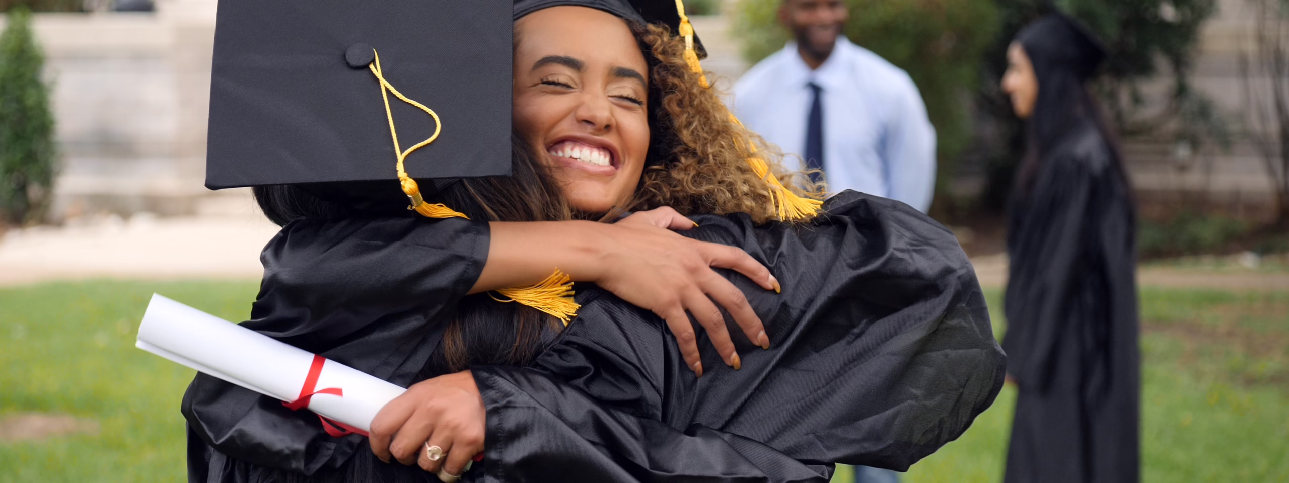 Two graduates in caps and gowns hug joyfully, one holding a diploma. Others in gowns are in the background