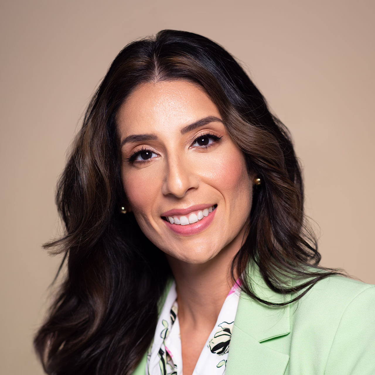 Smiling woman with long brown hair wearing a light green blazer and floral blouse against a beige background