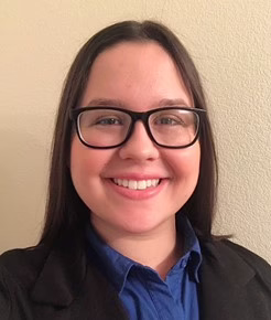 Smiling person with glasses and long dark hair, wearing a blue shirt and black jacket against a light background