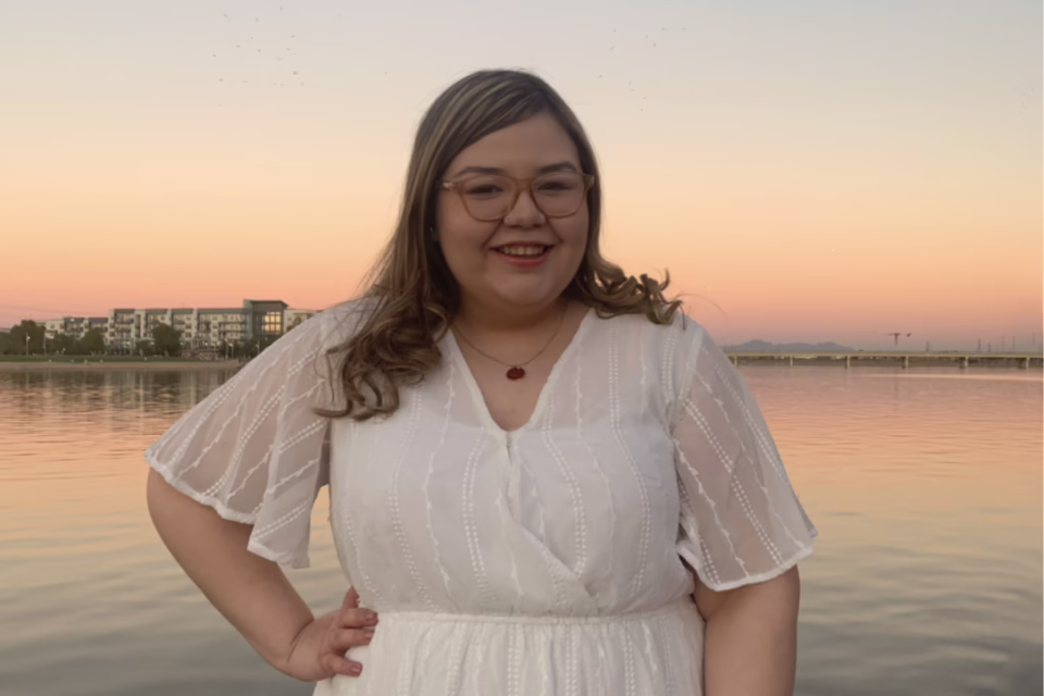 Person in a white dress smiling by a lake at sunset, with buildings in the background