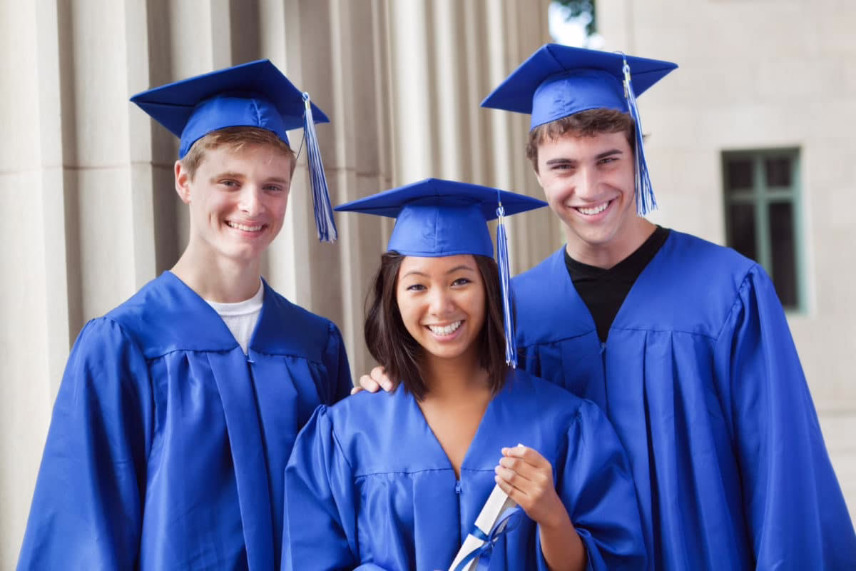 Three students in blue graduation gowns and caps smiling, one holding a diploma, standing in front of a building