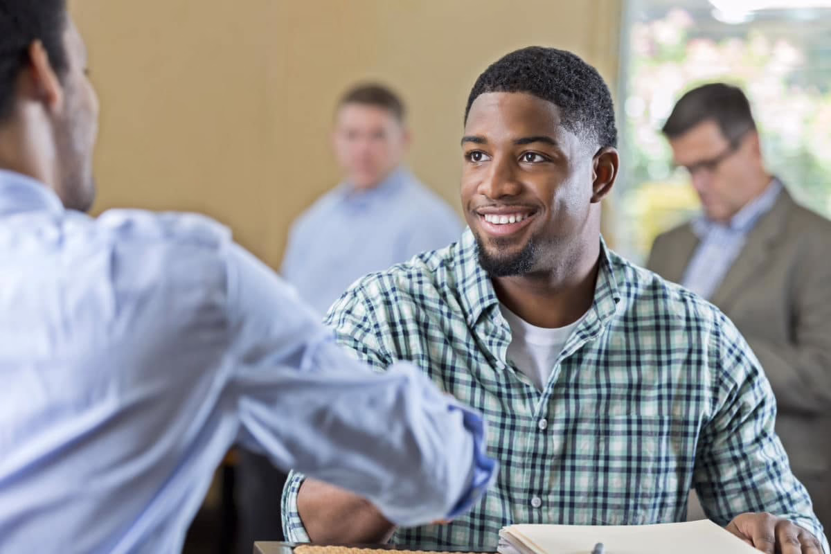 Young man in plaid shirt smiling and shaking hands with another person, with two men in the background
