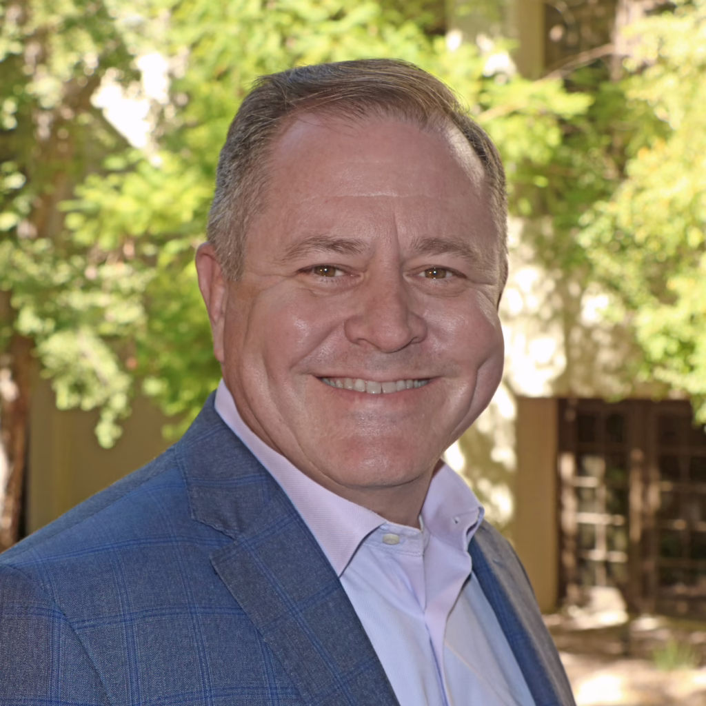 Smiling man in a blue suit jacket and white shirt, standing outdoors with trees in the background