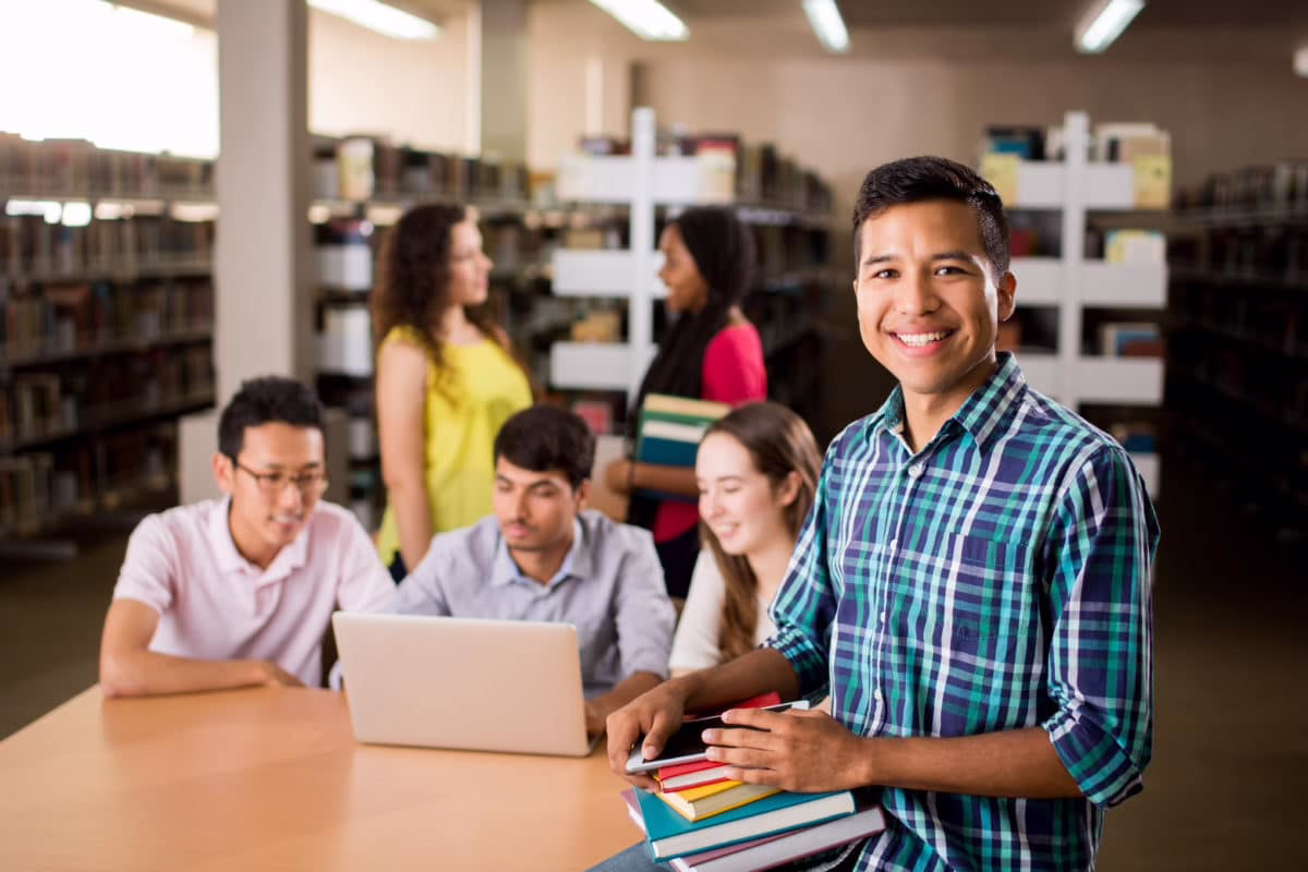 Students in a library, one smiling with books, others engaged with a laptop
