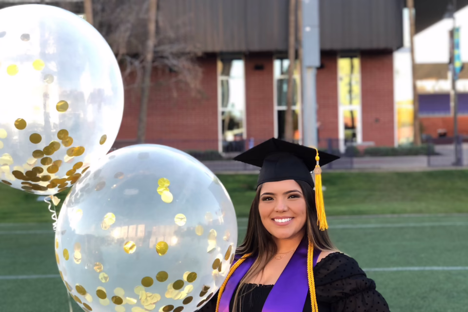 Graduate in cap and gown holding two large balloons with gold confetti, standing on grass with a building in the background