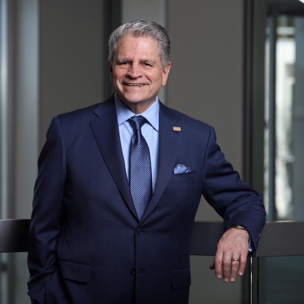 Man in a blue suit and tie, smiling, standing with one hand on a railing in a modern office setting