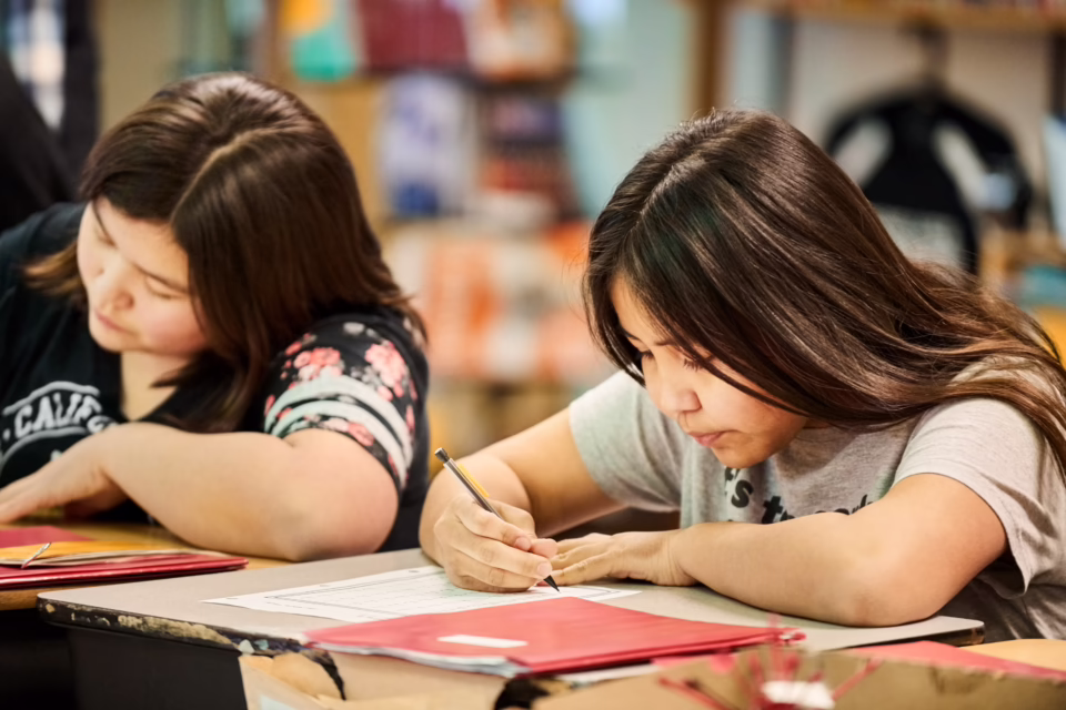 Two students focus on writing at their desks, surrounded by red folders and classroom materials
