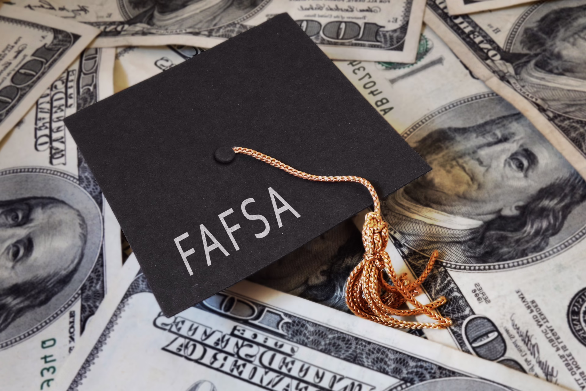 Graduation cap with "FAFSA" text on top of scattered hundred-dollar bills