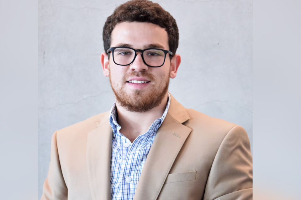 Man with glasses and a beard wearing a beige blazer and checkered shirt, smiling against a gray background