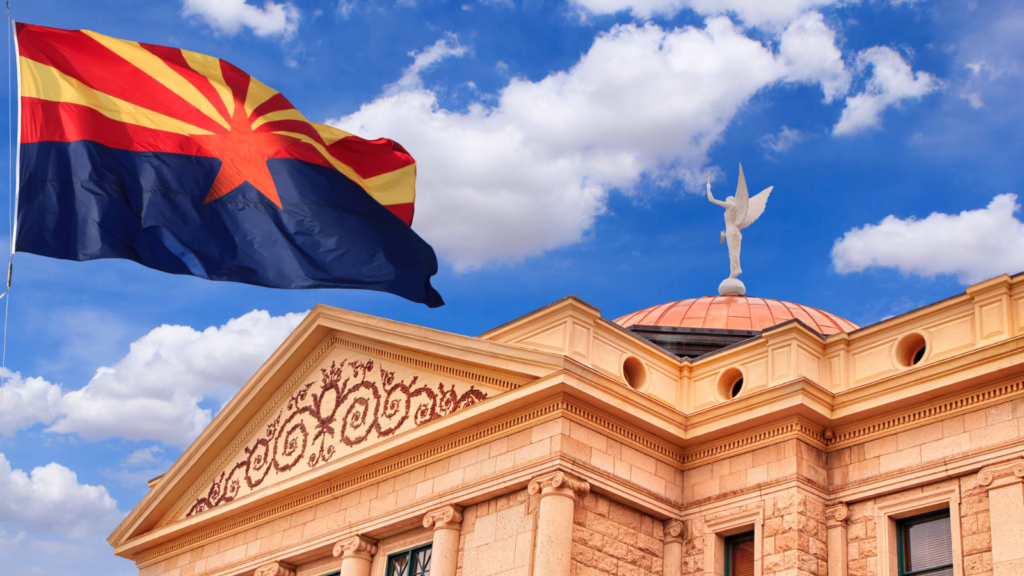 Arizona state flag waving near the Arizona Capitol building under a blue sky with clouds
