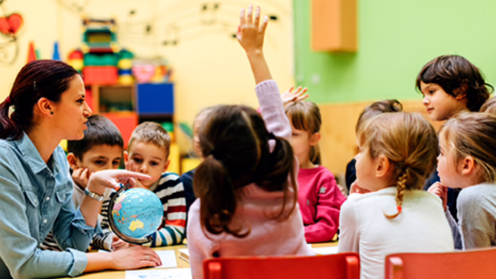 Teacher with globe engaging young students in a classroom; one child raises hand. Colorful decor in the background