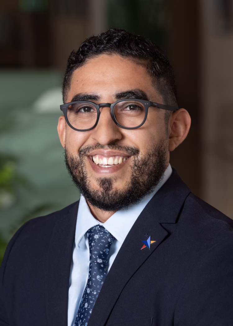 Smiling man with glasses and a beard, wearing a suit and tie, with a star pin on the lapel