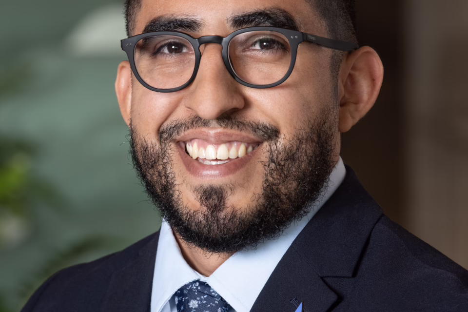Smiling man with glasses and a beard, wearing a suit and tie, with a star pin on the lapel