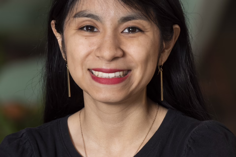 Smiling woman with long black hair, wearing a black top and gold earrings, in an indoor setting