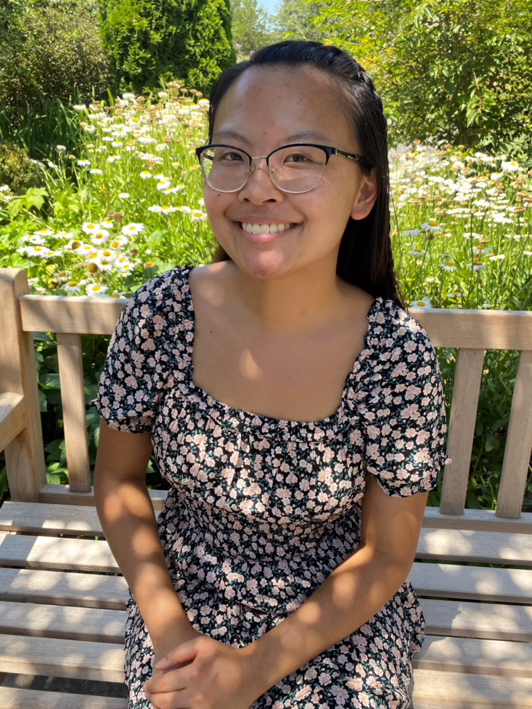 Smiling person with glasses sits on a bench in a garden with daisies, wearing a floral dress