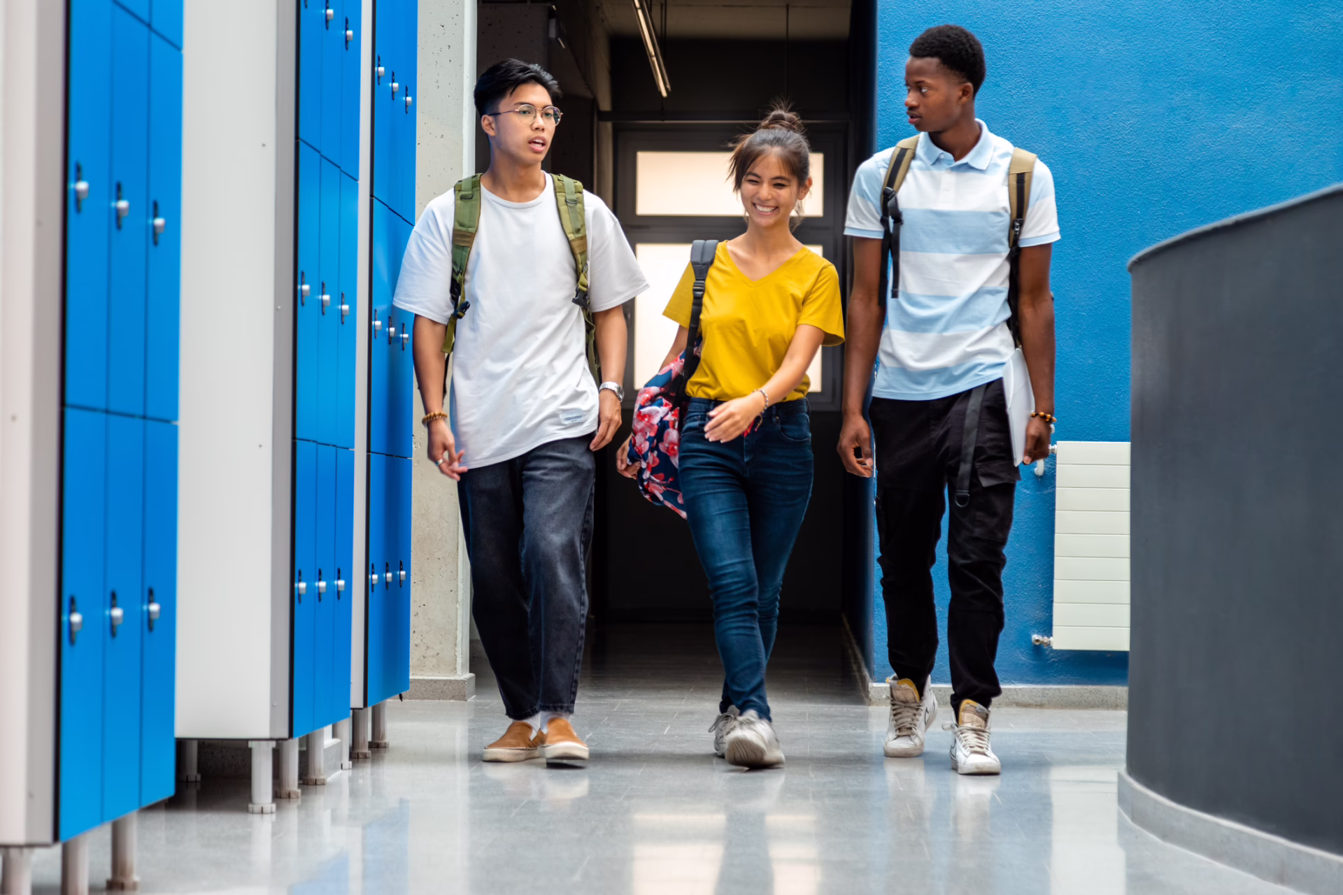 Three students with backpacks walk down a hallway with blue lockers, smiling and talking