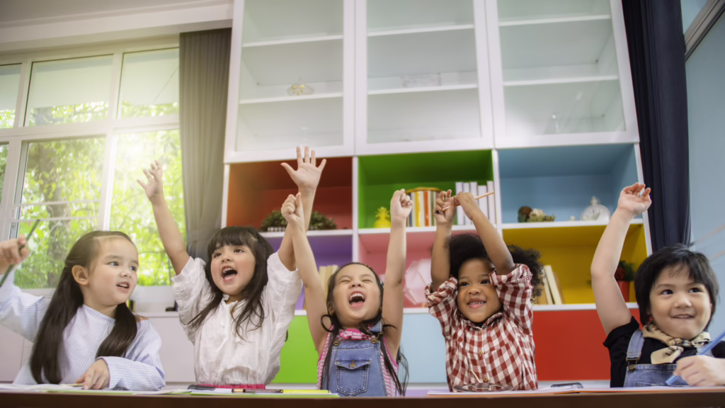 Children in a classroom with colorful shelves, raising their hands excitedly