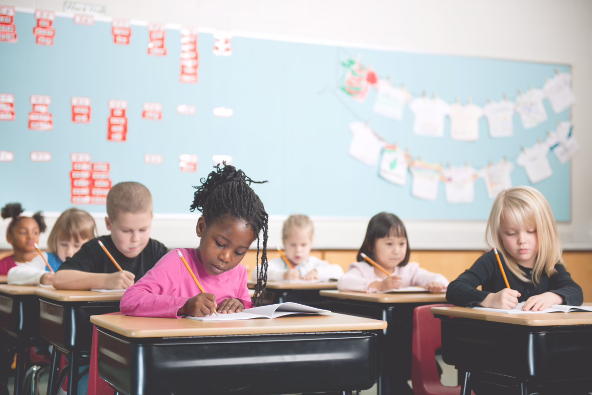 Children writing at desks in a classroom, with educational materials on the wall