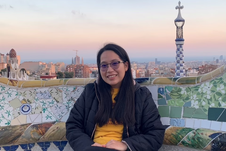 Smiling woman in a black jacket sits on a colorful mosaic bench with a cityscape and sunset in the background