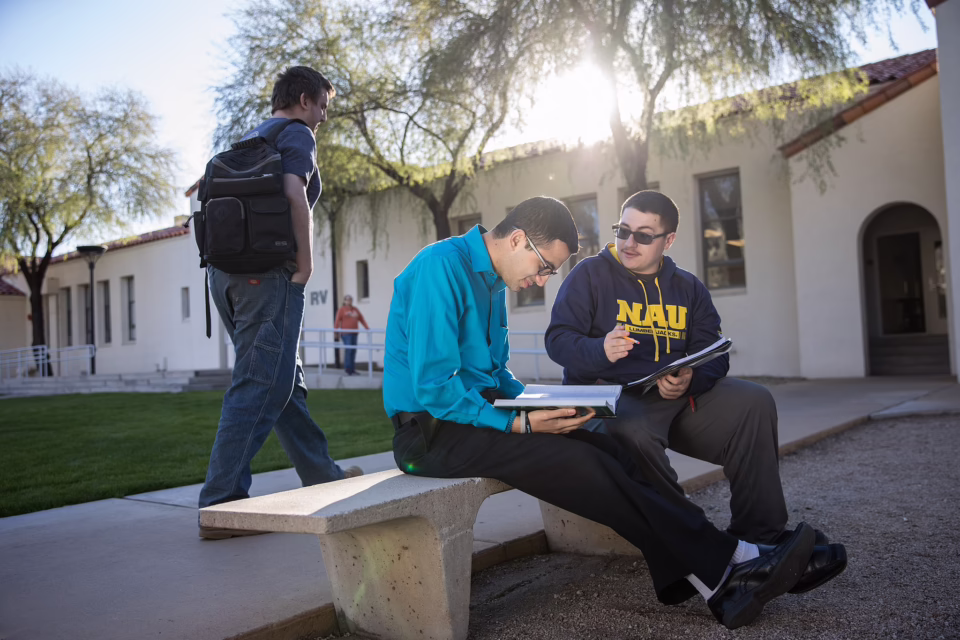 Two people sit on a bench with books, discussing, while another walks by on a sunny campus