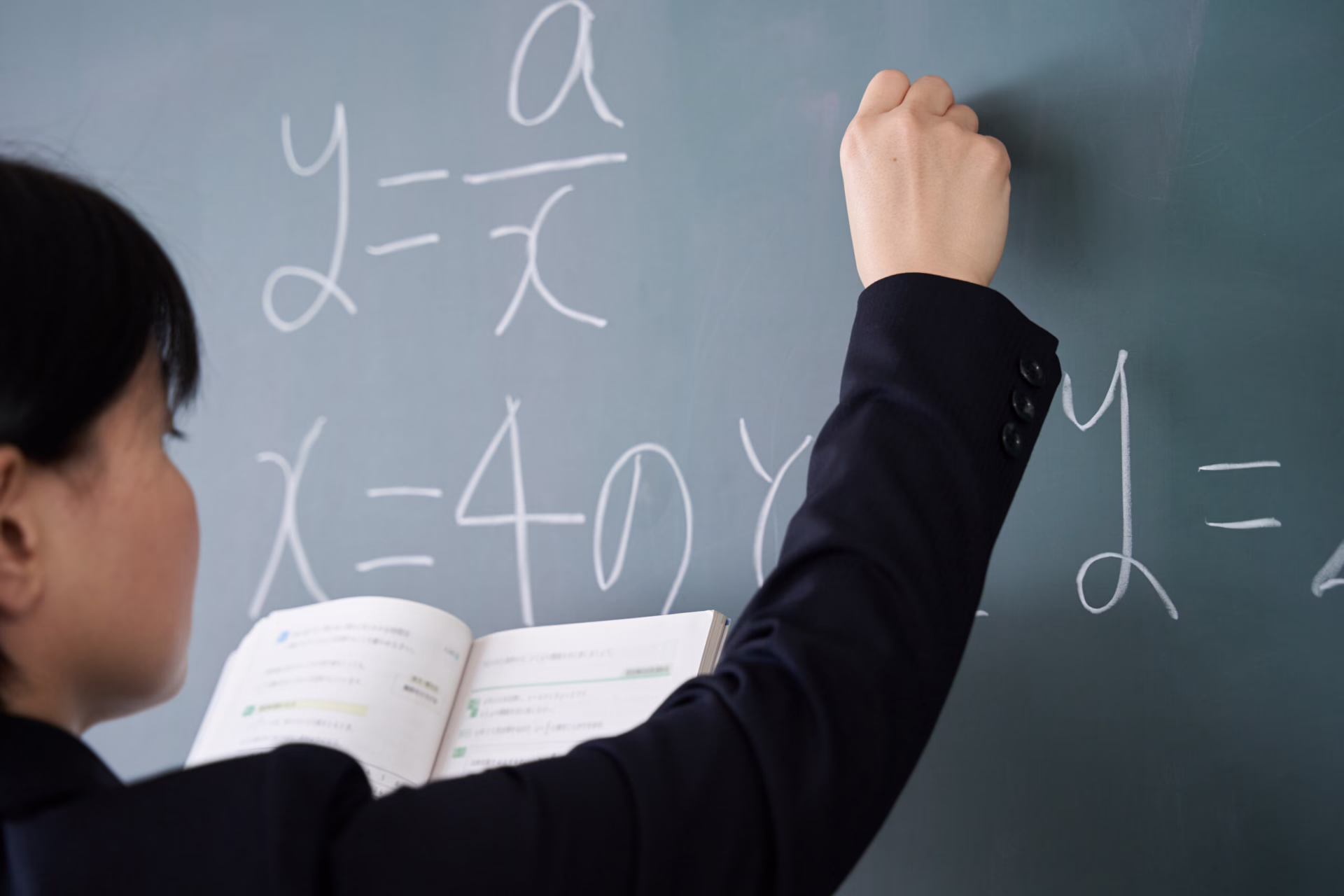 Person writing math equations on a chalkboard, holding an open book