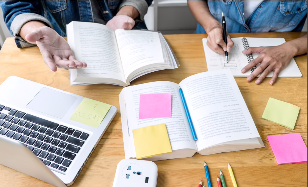 Open books, a laptop with "Lesson 1" sticky note, and notebooks on a desk. Two people studying and taking notes