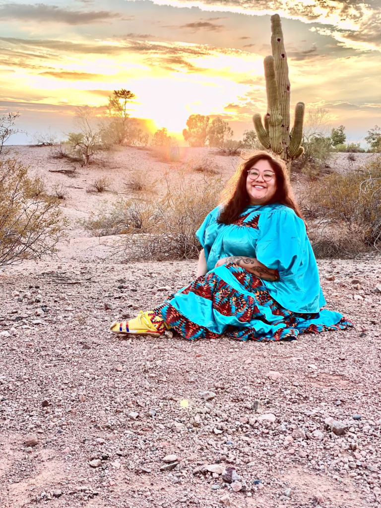 Woman in colorful attire sits in a desert landscape at sunset, with a cactus and distant trees in the background