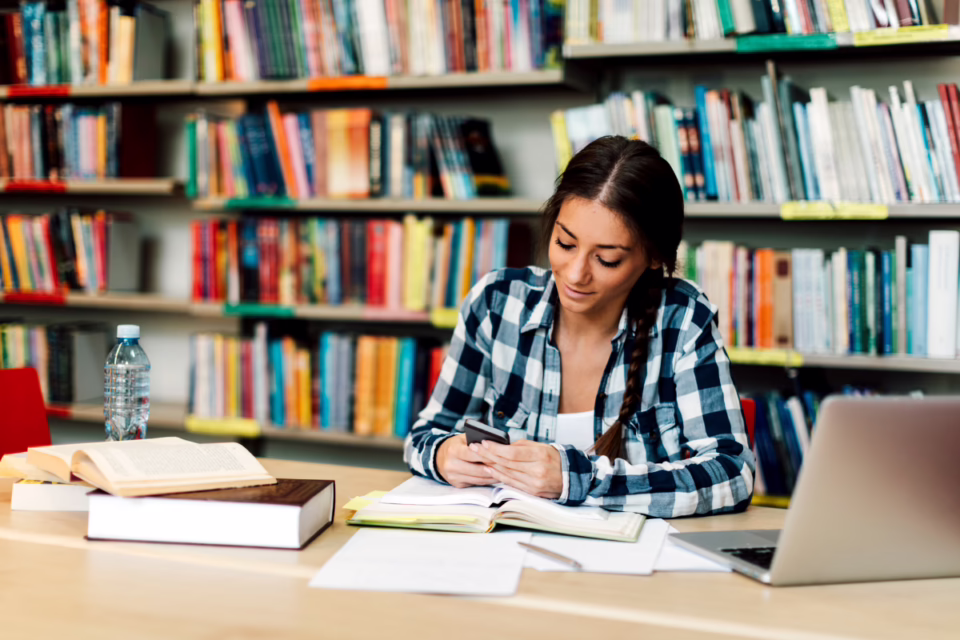 Student in a library using a smartphone, with books and a laptop on the table