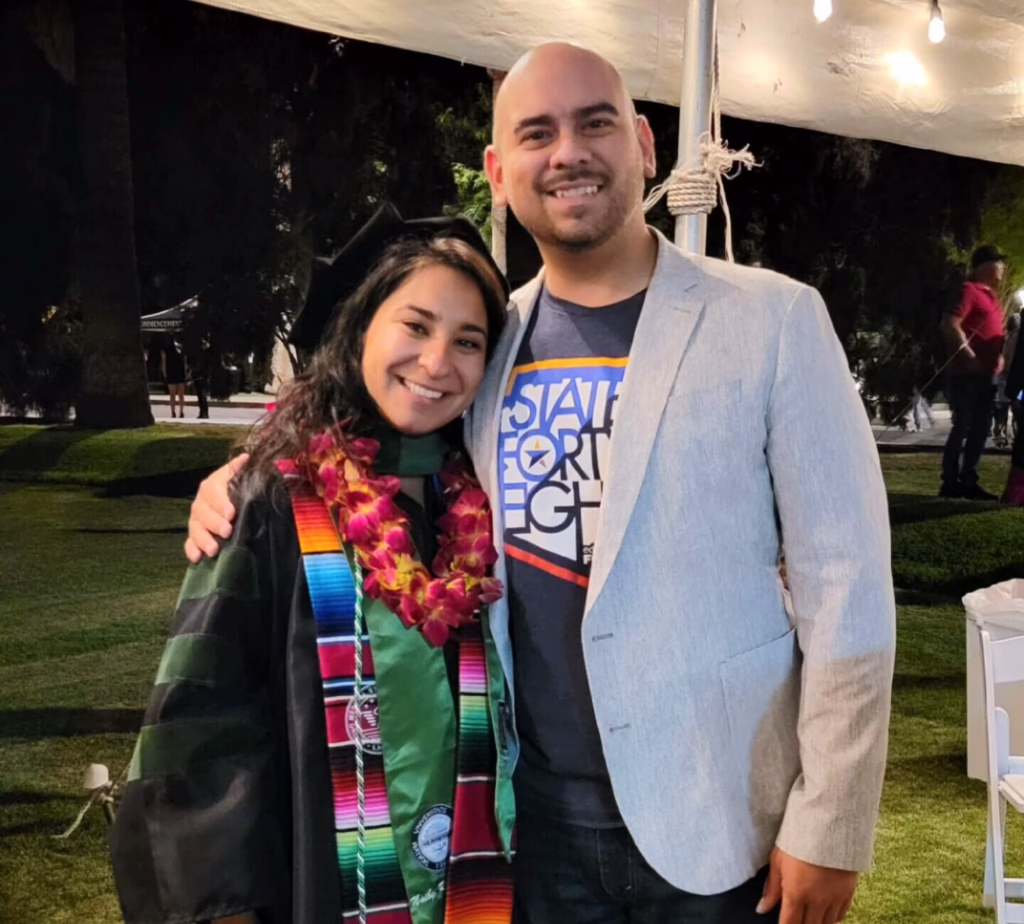 Graduate in cap and gown with colorful stole and lei, smiling with a man in a light blazer at an outdoor event