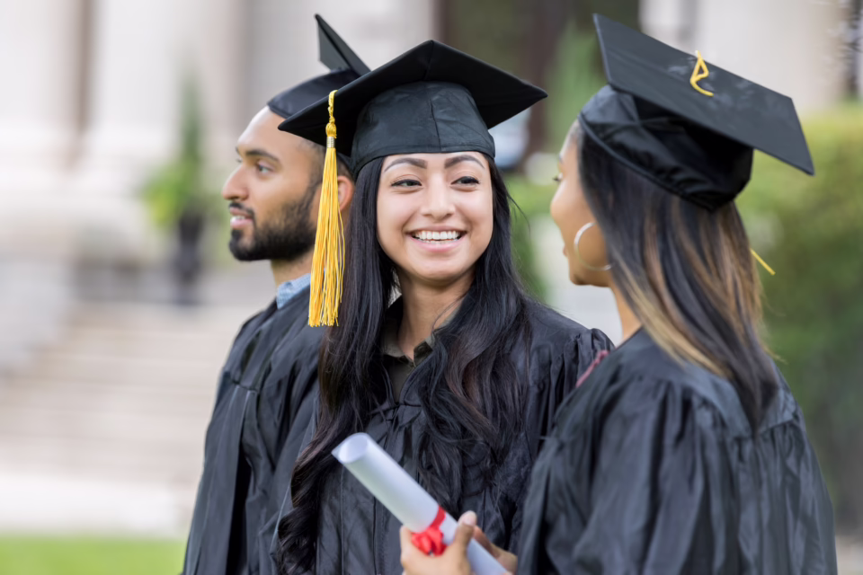 Three graduates in caps and gowns smile and chat, one holding a diploma