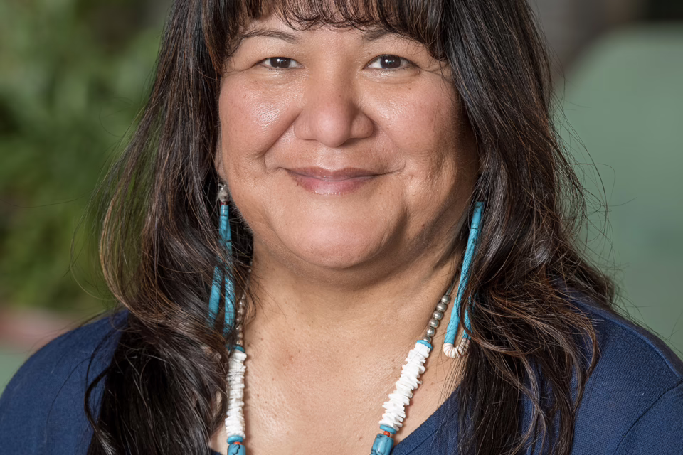 Smiling person with long dark hair, wearing a blue shirt and turquoise jewelry, standing in a blurred indoor setting
