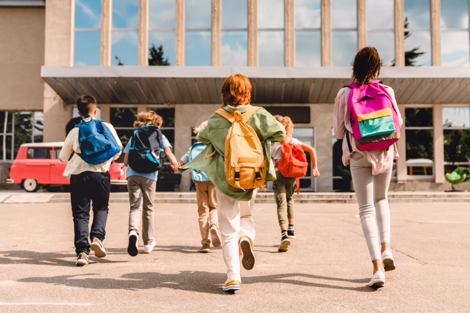 Children with colorful backpacks run towards a school building