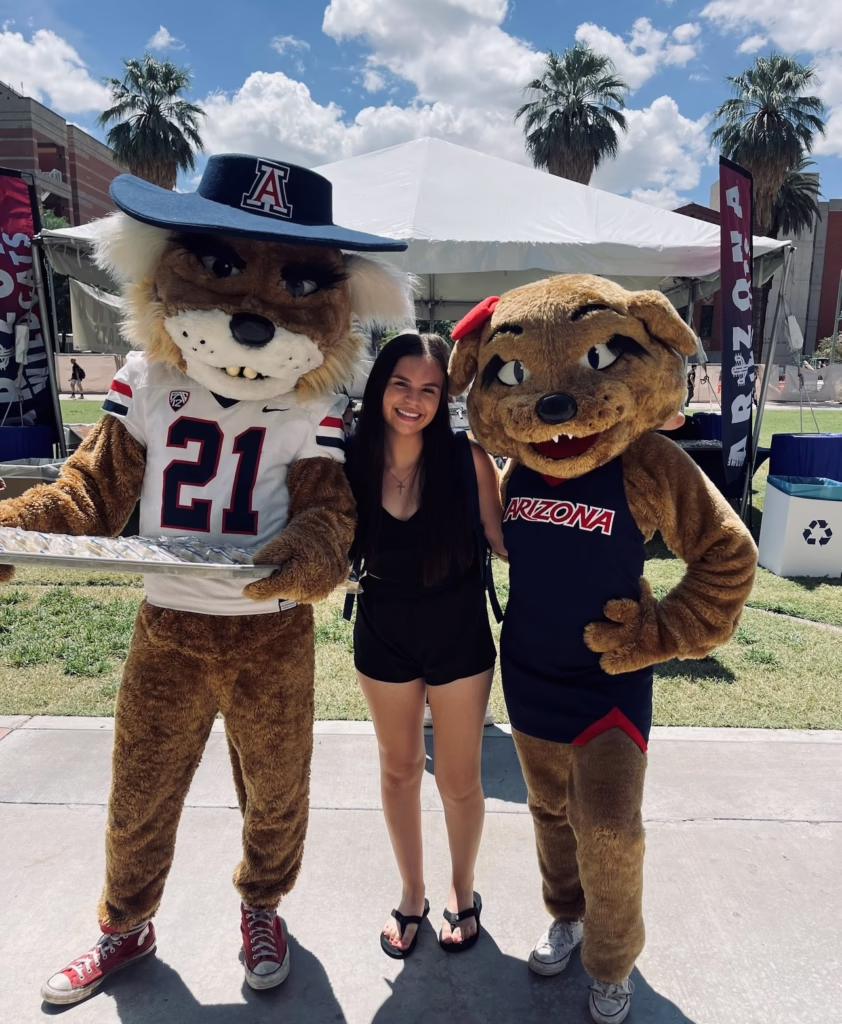 Student with Arizona mascots outdoors, palm trees and tent in background