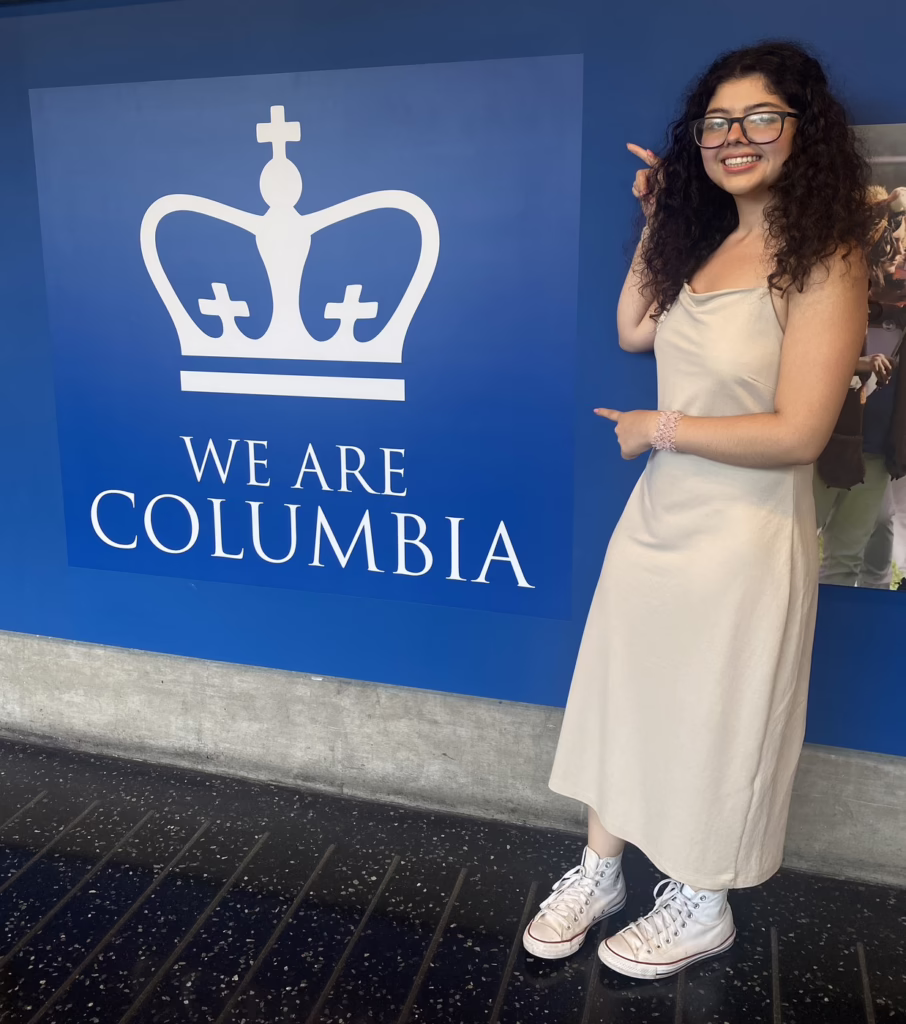 Person in a beige dress and sneakers smiles, pointing at a "We Are Columbia" sign with a crown logo