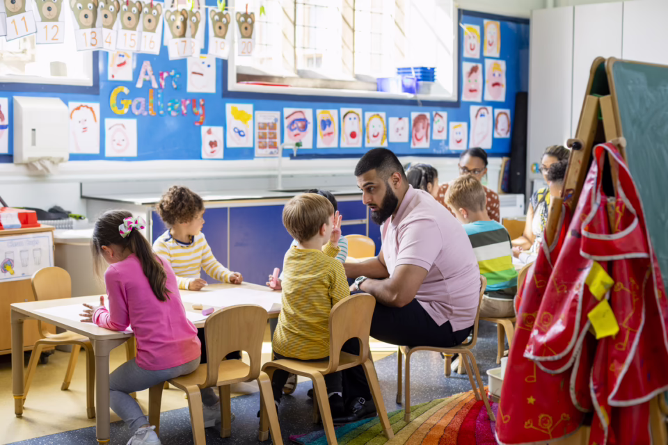 Children and a teacher in a classroom with art on the walls, discussing and drawing at a table