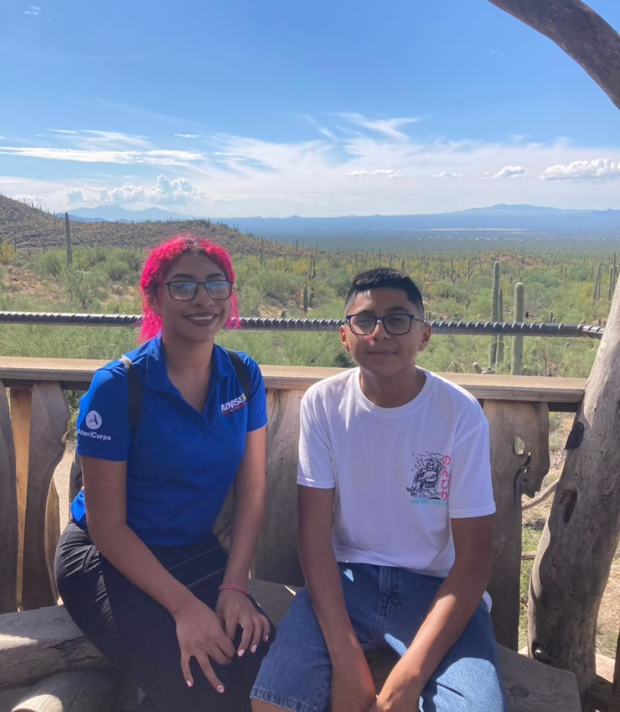 Two people sitting outdoors with a desert landscape in the background, one wearing a blue AdviseAZ shirt