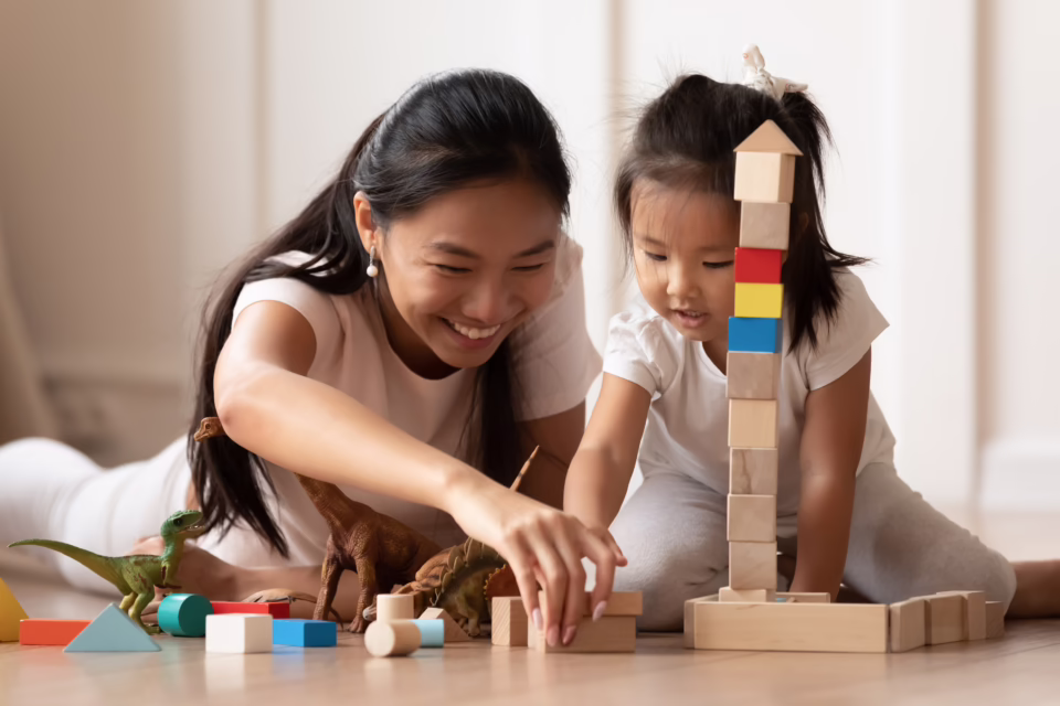 Mother and daughter playing with wooden blocks and toy dinosaurs on the floor, smiling and engaged