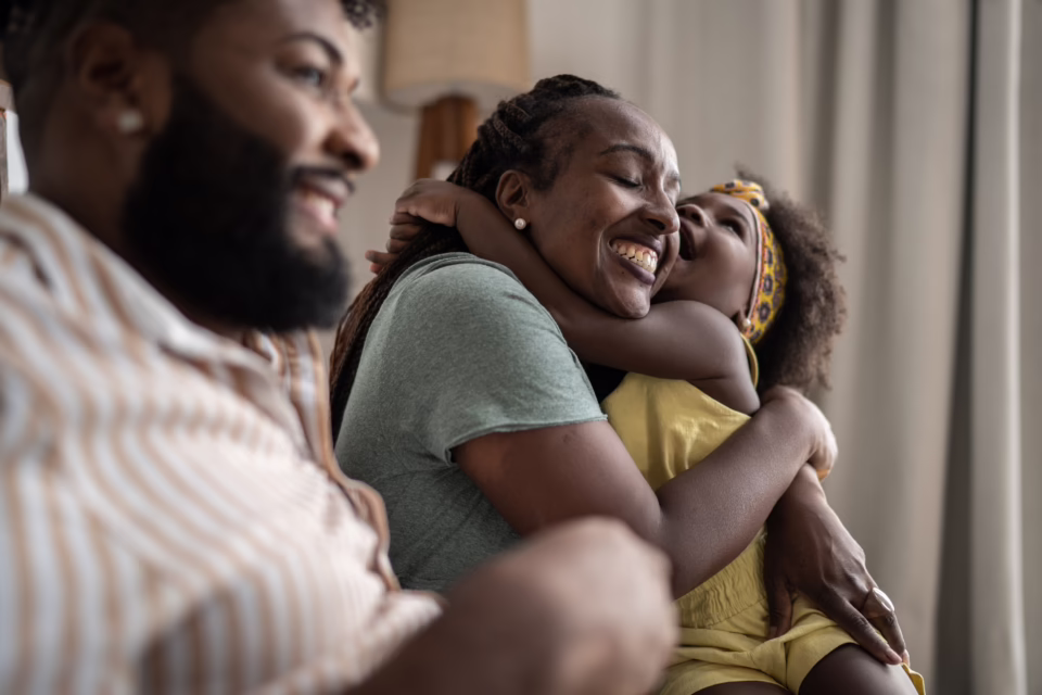 A joyful family moment with a child hugging an adult, both smiling, while another adult is in the foreground