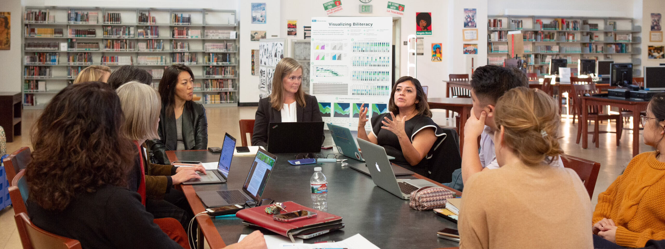 Group of people in a library discussing around a table with laptops and a "Visualizing Biliteracy" poster