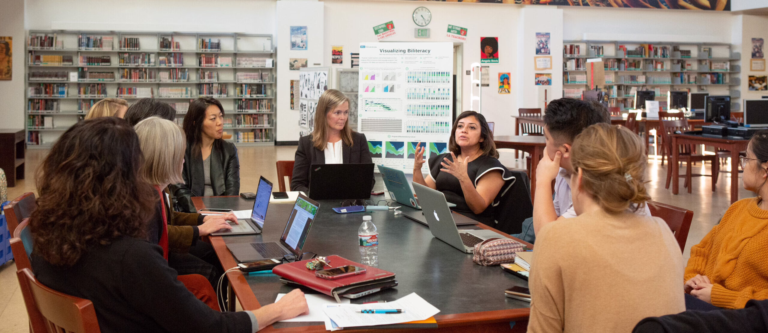 Group of people in a library discussing around a table with laptops and a "Visualizing Biliteracy" poster