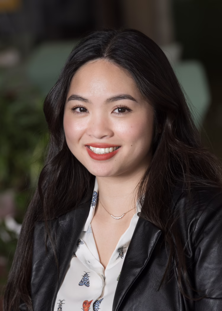 Smiling woman with long dark hair, wearing a patterned blouse and black jacket, in a blurred indoor setting