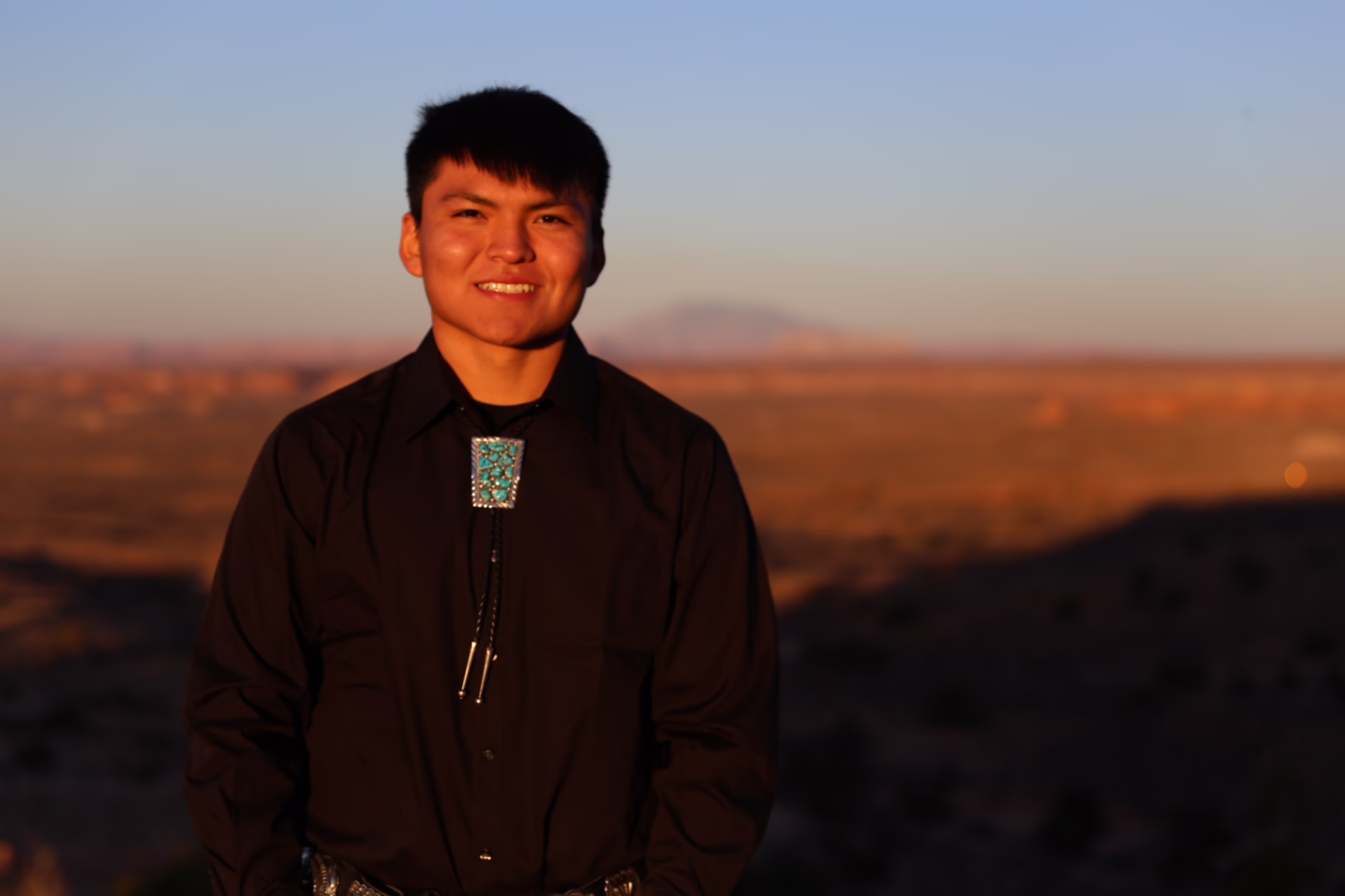Young man in a black shirt with a turquoise bolo tie, standing in a desert landscape at sunset