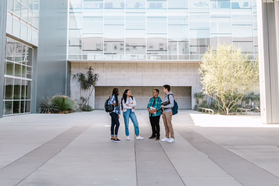 Four students with backpacks chat outside a modern college building