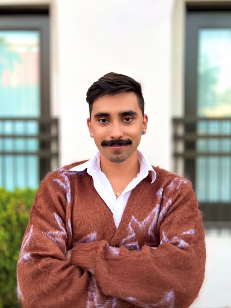 Person with a mustache wearing a brown sweater and white shirt, standing with arms crossed in front of a building