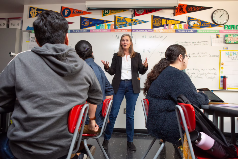 Teacher speaking to students in a classroom with college pennants and a whiteboard displaying notes